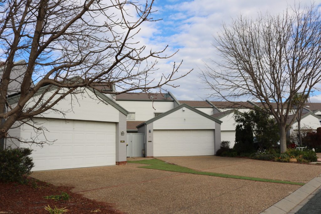 Exterior view of a strata unit complex in Greenway, Canberra, featuring multiple garages, landscaped gardens, and a cloudy sky, highlighting C&C Painting Group's exterior strata repaint project.