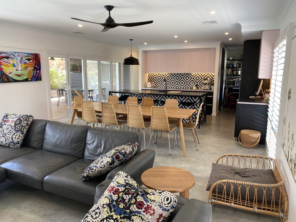 Modern living room and dining area showcasing stylish interior design, featuring a gray leather sofa, patterned cushions, a wooden coffee table, and a dining table with woven chairs, accented by colorful wall art and contemporary decor.