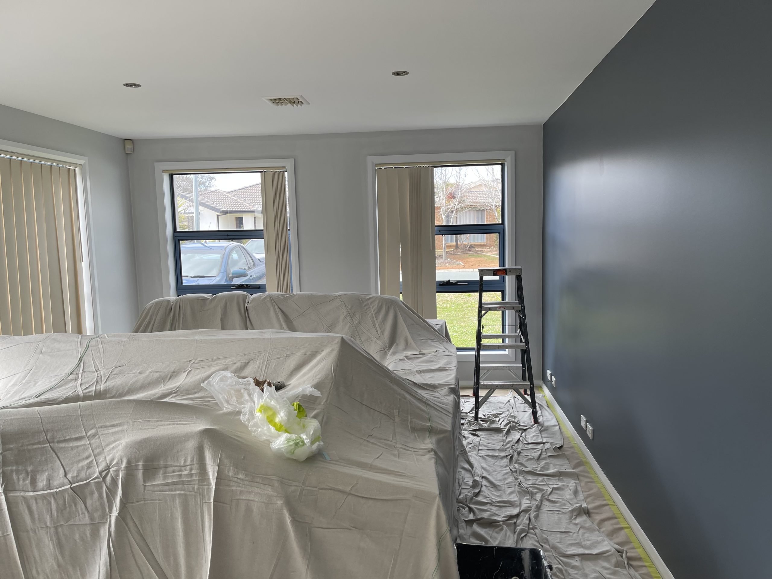 Interior room with covered furniture, a ladder, and freshly painted dark gray wall, showcasing preparation for painting, emphasizing professional interior house painting tips from C&C Painting Group.