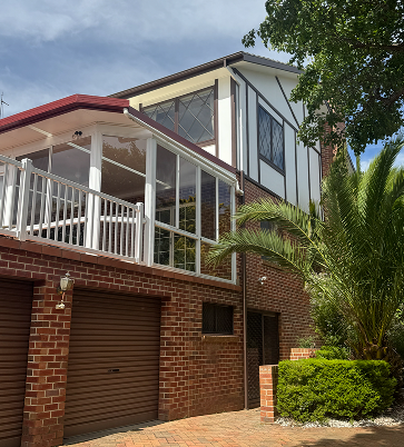Residential home exterior with modern architectural features, showcasing a repainted façade, large windows, and a balcony, surrounded by lush greenery and palm trees, reflecting C&C Painting Group's residential repainting services in Canberra.