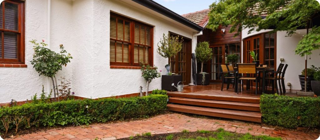 Exterior view of a residential home in Canberra featuring a well-maintained garden, wooden steps leading to a patio with outdoor dining furniture, and decorative plants alongside a white textured wall.