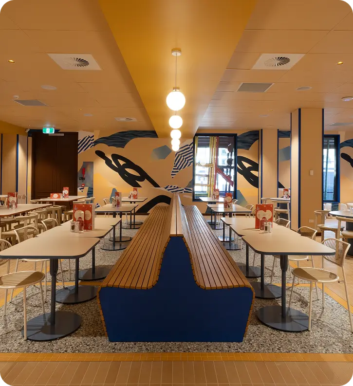 Modern dining area featuring wooden benches and tables, vibrant mural artwork on walls, pendant lighting, and a clean, contemporary design suitable for commercial spaces.