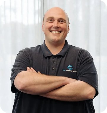 Smiling man with crossed arms in C&C Painting Group polo shirt, showcasing team spirit and professionalism, in an office setting.