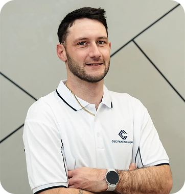 Portrait of a C&C Painting Group team member in a branded white polo shirt, smiling and standing confidently with arms crossed against a modern geometric background, representing the company's commitment to quality and professionalism in painting services.