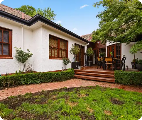Exterior view of a well-maintained home with a white facade, showcasing a landscaped garden, brick pathway, and wooden deck with outdoor seating, highlighting C&C Painting Group's residential repainting services in Canberra.