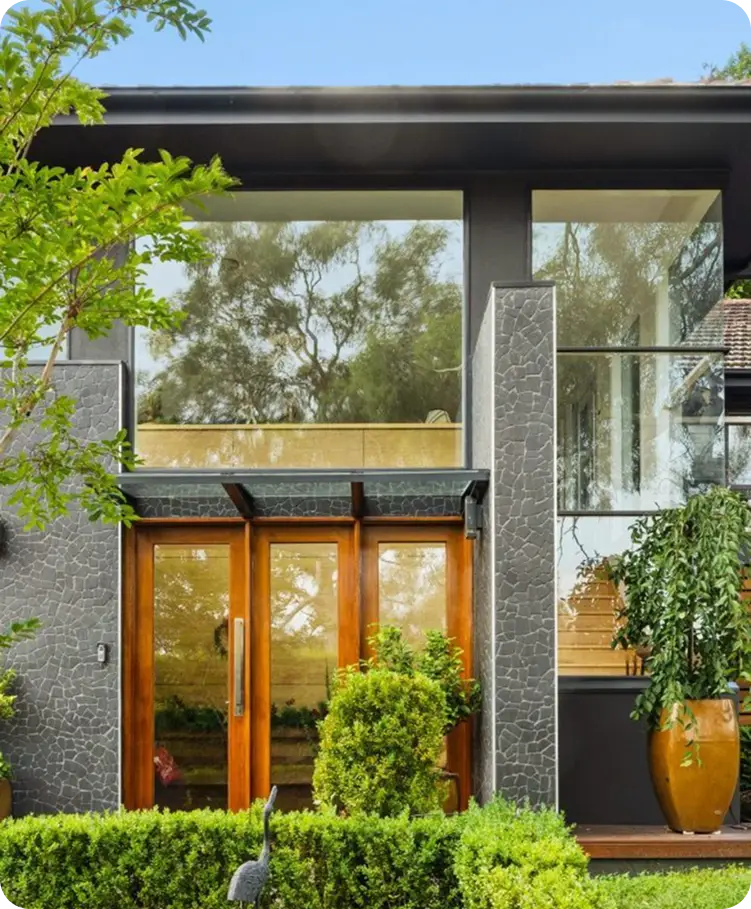 Modern home exterior with wooden double doors, large glass windows, and lush greenery, showcasing expert painting and landscaping by C&C Painting Group for residential transformations in Canberra.