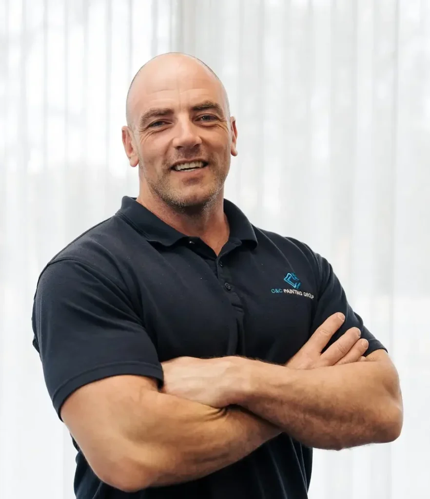 Luke Chamberlain, Co-Owner and Managing Director of C&C Painting Group, smiling with arms crossed, wearing a black polo shirt featuring the C&C Painting Group logo, against a backdrop of sheer curtains.