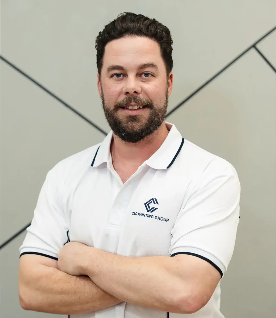 Adam McDonald, painter and team leader at C&C Painting Group, wearing a branded polo shirt with arms crossed, standing against a modern backdrop.